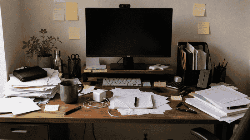 A cluttered home office desk covered in scattered papers, pens, cables, and random items representing a failed organization attempt