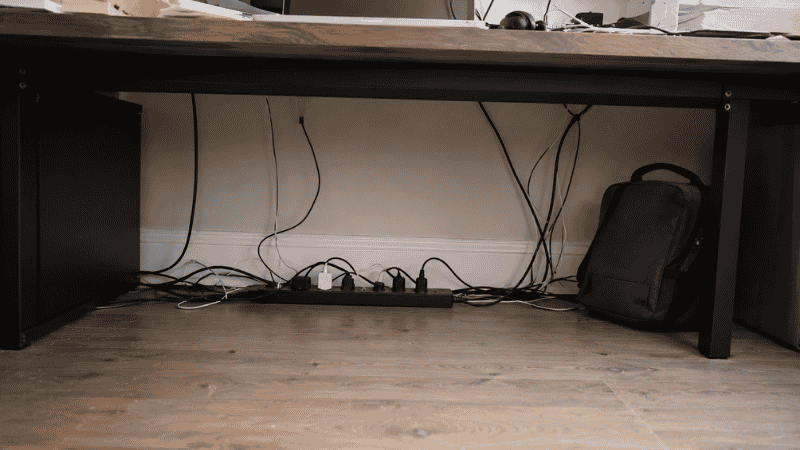 A cluttered under desk area with power strip on the floor, tangled cables, and unused space before organization