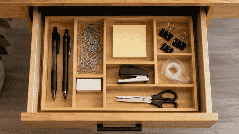 A well-organized desk drawer with compartment insert separating pens, paper clips, sticky notes, and scissors into neat assigned sections