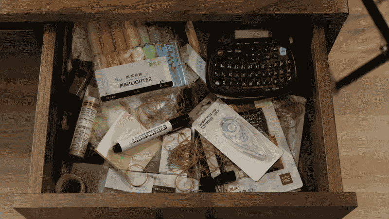 A label maker and other rarely used office supplies sitting unused in a cluttered desk drawer representing items that don't earn their space