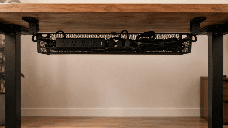 A metal mesh under desk cable management tray clamped beneath a desk holding a power strip and organized cables off the floor