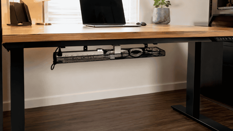 A metal mesh cable tray mounted under a desk holding a power strip and cables completely off the floor with the desk surface above looking clean