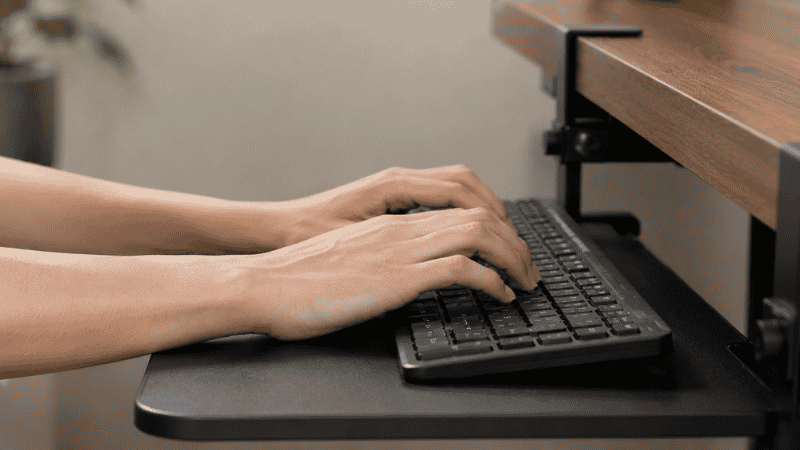 Hands on a keyboard mounted on an ergonomic keyboard tray below desk level with wrists in a neutral flat position for proper ergonomic typing