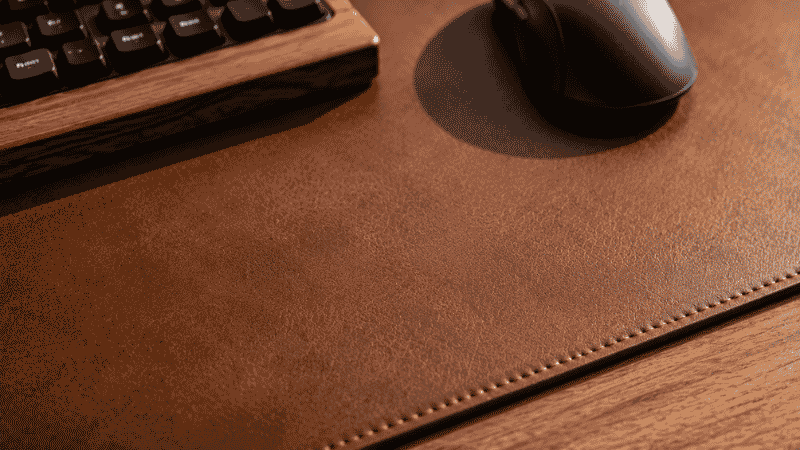 A close-up of a dark brown leather desk mat surface showing the smooth texture, stitched edges, and premium material quality