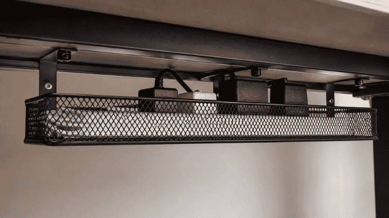 A metal mesh cable tray under a desk showing the open mesh construction with power adapters visible inside allowing airflow and ventilation