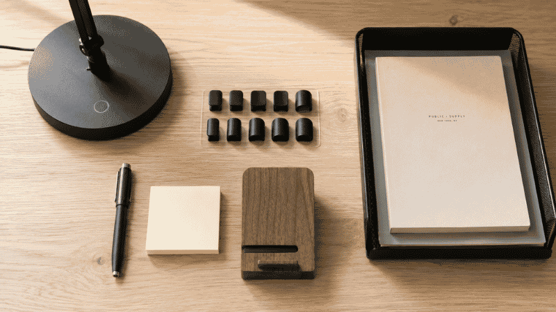 A flat lay of essential must-have desk accessories including cable clips, a pen, sticky notes, a small tray, and a phone stand arranged on a desk surface