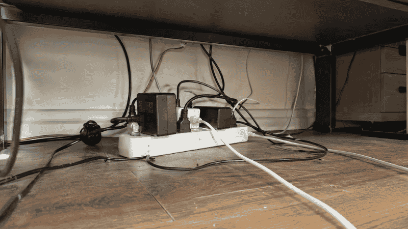 A power strip sitting on the floor under a desk with cables going in multiple directions showing the problem a cable tray solves