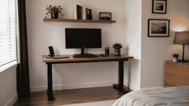 A small home office setup in a room corner with desk facing the wall, floating shelf above, and vertical storage making efficient use of limited space