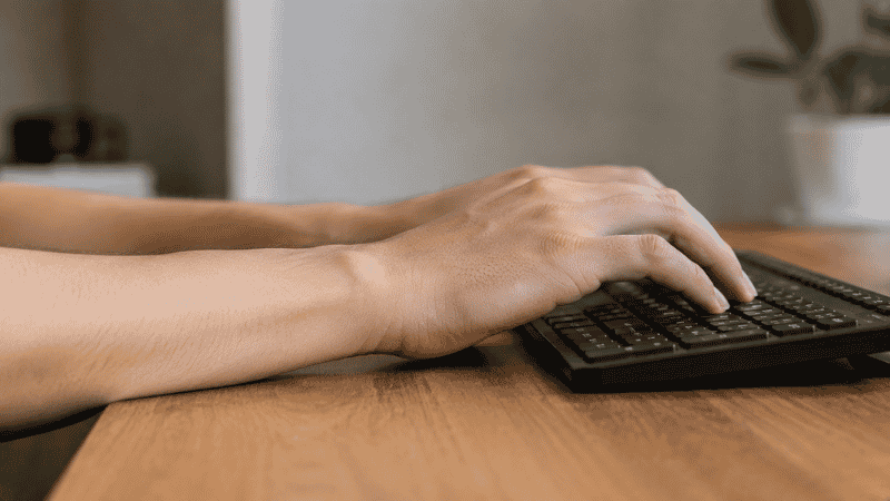 A close-up of hands on a keyboard at desk height showing wrists bent upward in an extended position indicating poor ergonomic positioning