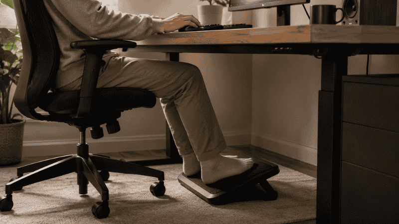 A footrest positioned under a home office desk supporting feet at the correct ergonomic height with the person seated at proper chair height