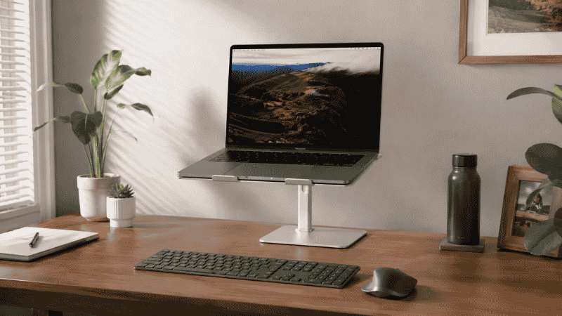 A laptop elevated on a stand at correct eye level on a home office desk with an external keyboard and mouse on the desk surface below