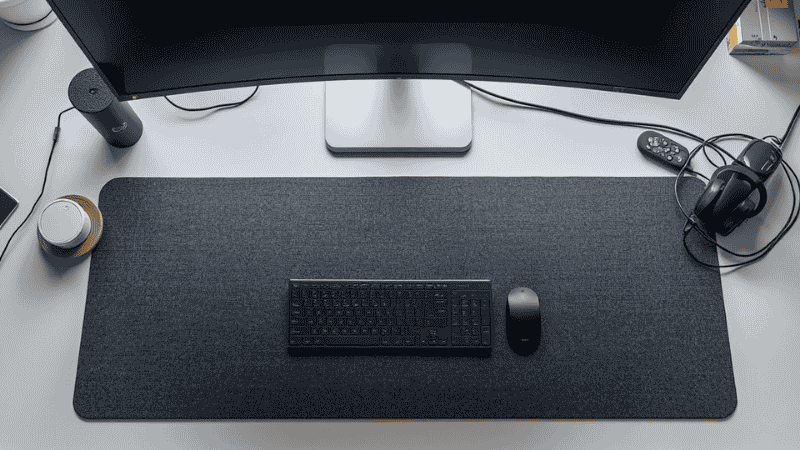 A top-down view of a modern 60-inch standing desk setup featuring an ultrawide monitor, a mechanical keyboard, and a large charcoal grey felt desk mat providing a tactile work surface.