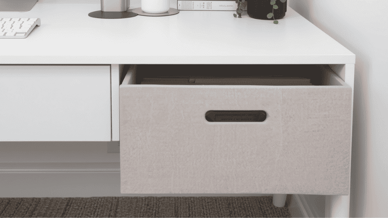 A clean, modern home office featuring a dual-monitor setup on a standing desk with a decorative grey linen file storage box organized on a nearby shelf for easy access.