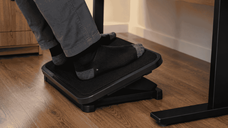 A standard padded footrest platform positioned under a desk with feet resting flat on its angled surface