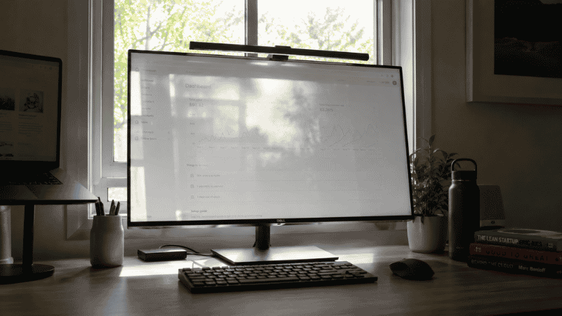 A home office desk with a window or bright light source behind the monitor creating glare and backlighting problems on the screen